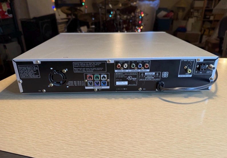 May include: Silver and black Sony DVD player with various input and output ports. The back panel features multiple connection options, including video, audio, and speaker ports. The device has a power cord and a fan for cooling.