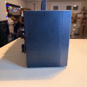 May include: A dark blue electronic device with a handle on top, resting on a light-colored wooden surface. The device has a rectangular shape with a few visible buttons and a power cord. The background is blurred.