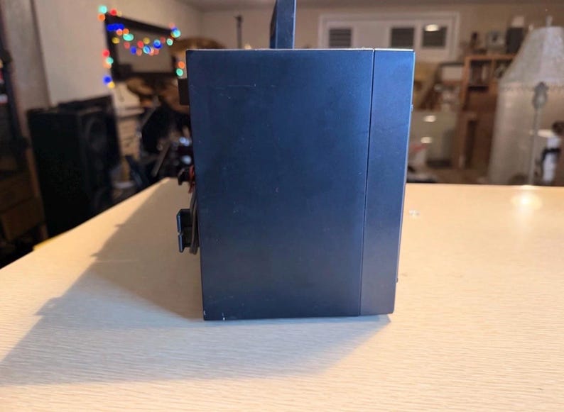 May include: A dark blue electronic device with a handle on top, resting on a light-colored wooden surface. The device has a rectangular shape with a few visible buttons and a power cord. The background is blurred.