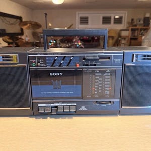 May include: A vintage Sony CFS-9000 boombox, featuring a cassette player, AM/FM radio, and graphic equalizer. The unit is primarily gray with black accents, a carrying handle, and speaker grilles on each side. The radio dial displays FM and AM frequencies.