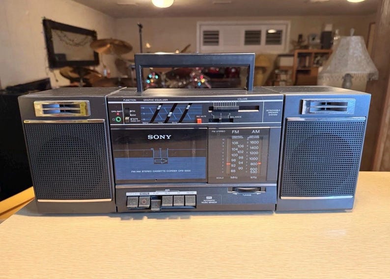 May include: A vintage Sony CFS-9000 boombox, featuring a cassette player, AM/FM radio, and graphic equalizer. The unit is primarily gray with black accents, a carrying handle, and speaker grilles on each side. The radio dial displays FM and AM frequencies.