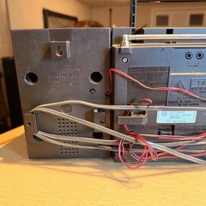 May include: A black Sony boombox with a silver antenna and red and gray wires. The boombox has a speaker on the left side and a control panel on the right. The boombox is sitting on a light brown surface.