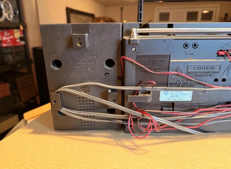 May include: A black Sony boombox with a silver antenna and red and gray wires. The boombox has a speaker on the left side and a control panel on the right. The boombox is sitting on a light brown surface.