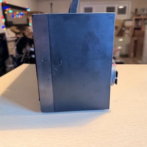 May include: A dark blue, rectangular electronic device with a handle and a dial on the side. The device is sitting on a light-colored wooden surface. The background is blurred.