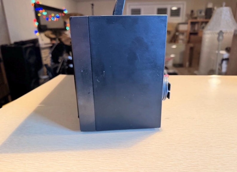 May include: A dark blue, rectangular electronic device with a handle and a dial on the side. The device is sitting on a light-colored wooden surface. The background is blurred.