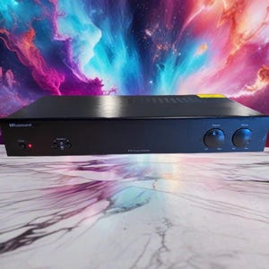 Puede incluir: Un amplificador de potencia negro Russound P75 con un panel frontal que incluye un botón de encendido, una luz indicadora roja, dos botones de selección de altavoces y dos perillas circulares para ajustar el balance y el volumen.