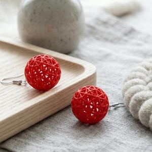 May include: Two red, spherical earrings with an open, geometric design. The earrings are displayed on a light wooden tray, with a grey fabric background. The earrings have a hook closure.