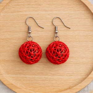 Può includere: Un paio di orecchini pendenti con ciondoli sferici rossi caratterizzati da un intricato motivo geometrico a reticolo traforato. Ogni sfera è sospesa a un gancio in metallo color argento, creando un design leggero e pendente.
