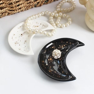 May include: Two crescent-shaped trinket dishes. One is white with gold speckles, and the other is black with gold speckles and a white flower accent. A pearl necklace rests on the white dish.