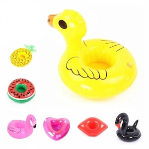 Op de afbeelding: Een verzameling van zeven opblaasbare bekerhouders in diverse vormen en kleuren, waaronder een gele eend, een ananas, een watermeloen, een roze flamingo, een roze hart, rode lippen en een zwarte zwaan, allemaal ontworpen om op water te drijven.