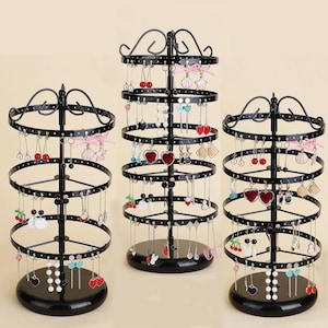 Peut inclure: Trois présentoirs à bijoux rotatifs en métal noir avec plusieurs niveaux circulaires pour suspendre des boucles d'oreilles. Chaque présentoir possède un mât central, une base ronde et des volutes décoratives, présentant diverses boucles d'oreilles.