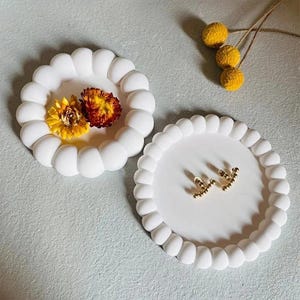 Può includere: Due vassoi decorativi rotondi bianchi con bordo perlato. Un vassoio contiene due fiori secchi, l'altro un paio di orecchini dorati. I vassoi sono su una superficie grigio chiaro. Ideali per gioielli.