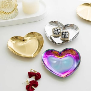 May include: A collection of heart-shaped trinket dishes in gold, silver, and iridescent finishes. One silver dish holds square earrings with black and white patterns. A pair of red bow earrings are also visible.