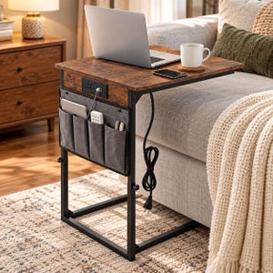 May include: A C-shaped side table with a dark wood-grain top, a black metal frame, and a built-in power outlet. It features a gray fabric storage pocket on the side containing a remote and pens, with a power cord hanging from the frame.