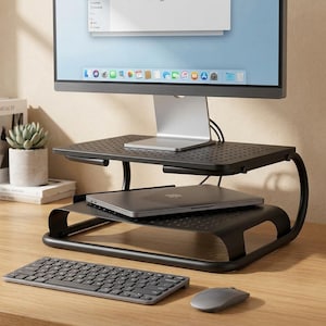 May include: A black metal two-tier monitor stand with a perforated surface design. The stand supports a computer monitor on the top shelf and a closed laptop on the lower shelf. A wireless keyboard and a computer mouse are placed in front of the stand.