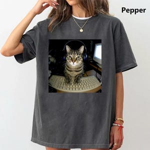 May include: A charcoal gray t-shirt featuring a graphic print of a tabby cat wearing headphones while sitting at a computer keyboard. The shirt has a crew neckline and short sleeves. The word Pepper is printed in the upper right corner of the image.