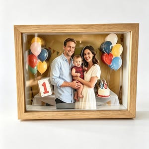 May include: A light oak shadow box frame containing a three-dimensional layered photograph of two adults holding a baby. The scene features colorful balloons, a white cake with a number one topper, and a sign with the number one in red.