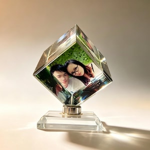 May include: A clear glass cube photo display featuring a color photograph of two people embedded inside. The cube is mounted at an angle on a square glass base with a small metallic connector, creating a reflective and transparent display piece.