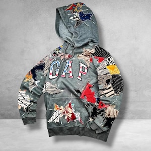 May include: A grey hooded sweatshirt featuring the text GAP in white letters with a red outline. The garment is decorated with various colorful fabric patches in floral, striped, and solid patterns, stitched onto the sleeves, hood, and front.