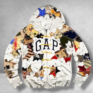 May include: A light gray zip-up hoodie featuring the navy blue text GAP on the chest. The garment is decorated with numerous multicolored fabric patches in various patterns and shapes, attached with visible white stitching and yellow toggle closures.