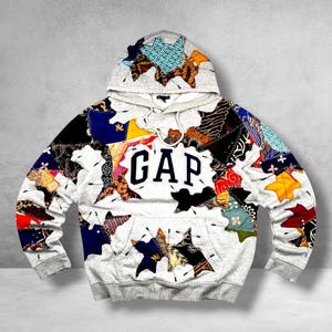 May include: A light gray hooded sweatshirt featuring a prominent navy blue GAP logo on the chest. The garment is decorated with numerous multicolored fabric patches in various shapes and patterns, all secured with visible dark stitching throughout.