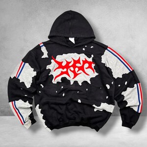 May include: A black hooded sweatshirt featuring a large red graphic on the chest, irregular white patches across the front and sleeves, and vertical red, white, and blue stripes running down the outer length of each sleeve.