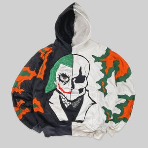 Puede incluir: Una sudadera con capucha de diseño dividido con un gráfico de medio Joker y medio calavera. La sudadera es negra y blanca con detalles en naranja y verde. La capucha está dividida en negro y blanco. El diseño incluye una ilustración detallada de la cara del Joker y una calavera.