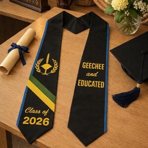 Może przedstawiać: Czarna szarfa absolwencka z niebieskim wykończeniem, z napisem "Geechee and Educated" i "Class of 2026". Szarfa ma złoty i zielony emblemat. Widoczna jest również czarna czapka absolwencka z niebieskim chwostem.