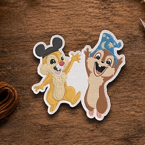 May include: An embroidered patch featuring two cartoon chipmunks. The left chipmunk wears black mouse ears, while the right chipmunk wears a blue wizard hat with yellow stars and moons. Both figures have raised arms and wide, happy expressions.