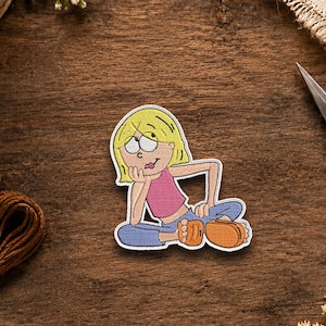 May include: A fabric patch featuring a cartoon illustration of a blonde character with a pink top and blue pants, sitting with one hand resting on their chin. The character has large eyes and is wearing orange footwear, rendered in a flat graphic style.