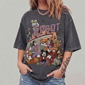 May include: A charcoal grey short-sleeved t-shirt featuring a graphic print with the text EPCOT in purple letters. The design includes cartoon characters dressed in Halloween costumes, pumpkins, and a monorail track against a moonlit background.