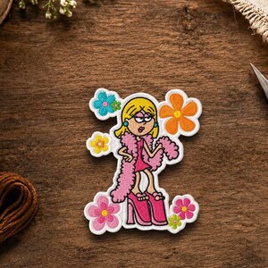 May include: An embroidered patch featuring a cartoon character with blonde hair wearing a pink dress, a pink feather boa, and pink platform shoes, surrounded by colorful embroidered flowers in shades of blue, orange, yellow, and pink.