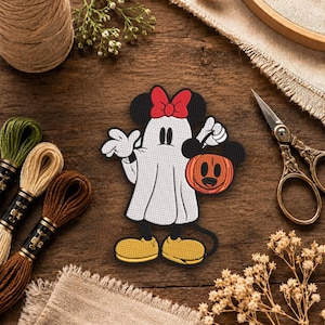 May include: An embroidered patch featuring a character dressed as a white ghost with a red bow, wearing yellow shoes, and holding an orange jack-o'-lantern bucket with a black mouse-ear silhouette design on the front of the pumpkin.
