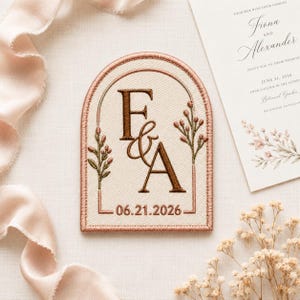 May include: An arched embroidered patch featuring the letters F and A in dark brown, an ampersand, and the date 06.21.2026. The design includes a copper-colored border and two floral sprigs with green stems and small pinkish-brown buds.