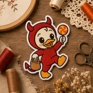 May include: An embroidered patch featuring a cartoon duckling wearing a red devil costume with horns and a tail, holding a brown bucket in one hand and an orange and yellow swirled lollipop in the other, outlined in white on a dark background.