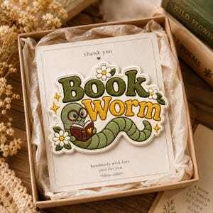May include: An embroidered patch featuring a green worm wearing glasses and reading a brown book, with the text "BOOK WORM" in green and yellow. The patch is decorated with white daisies and yellow stars, presented on a card inside a gift box.