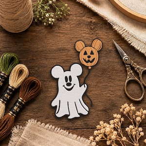 May include: An embroidered patch featuring a white ghost character with round ears holding a black string attached to an orange pumpkin balloon with a jack-o'-lantern face and matching round ears, outlined in black thread on a white background.