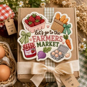 May include: An embroidered patch featuring the text 'let's go to the FARMERS MARKET' surrounded by illustrations of a strawberry basket, bread, a jam jar, a purple turnip, a carrot, and an egg carton, centered on a brown gift box with a ribbon.