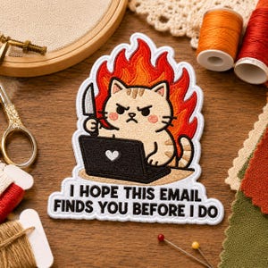 May include: An embroidered patch featuring a beige cat with a grumpy expression, holding a knife while sitting at a black laptop with a white heart icon. The cat is surrounded by orange and red flames. The text reads, I HOPE THIS EMAIL FINDS YOU BEFORE I DO.