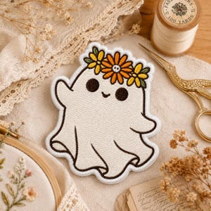 May include: An embroidered patch featuring a white ghost with a smiling face and dark eyes, wearing a crown of yellow and orange flowers. The patch has a thick white border and is stitched with detailed thread work, showcasing a playful design.