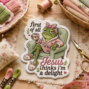 May include: An embroidered patch featuring a green frog wearing a pink robe, headband, and bunny slippers, sitting in a green armchair while holding a mug. The text reads: first of all Jesus thinks i'm a delight, with small heart accents.