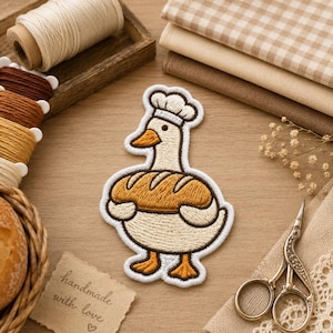 May include: An embroidered patch featuring a white duck wearing a chef hat and holding a golden-brown loaf of bread. The duck has orange feet and an orange beak, with dark brown outlines defining its shape against a light-colored background.