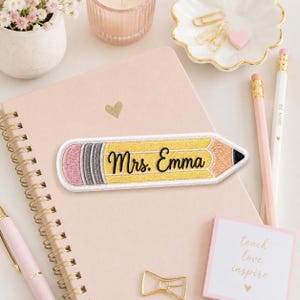 May include: A pencil-shaped embroidered patch featuring a yellow body, a pink eraser, a grey ferrule, and a tan tip. The text "Mrs. Emma" is stitched in black cursive script across the center of the yellow pencil body with a white border.