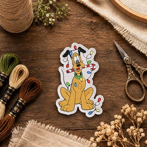 May include: An embroidered patch featuring a yellow cartoon dog with black ears, wearing a green collar, holding a red and white striped candy cane in its mouth, and surrounded by a string of colorful Christmas lights on a white background.