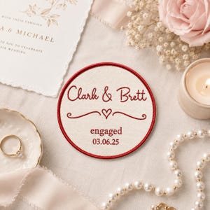 May include: A circular fabric patch with a red embroidered border on a cream background. The text reads Clark & Brett, followed by a small heart icon, the word engaged, and the date 03.06.25, all stitched in a matching red thread.