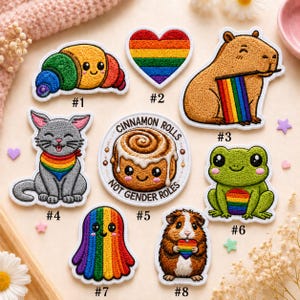 May include: A collection of eight embroidered patches featuring a rainbow croissant, a rainbow heart, a capybara with a rainbow flag, a gray cat with a rainbow bandana, a cinnamon roll with the text CINNAMON ROLLS NOT GENDER ROLES, a green frog, a rainbow ghost, and a guinea pig holding a rainbow heart.