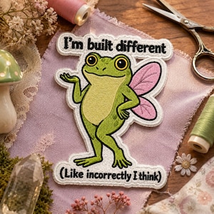 May include: An embroidered patch featuring a green frog with pink wings, standing with one hand on its hip. The text reads "I'm built different" at the top and "(Like incorrectly I think)" at the bottom, all outlined in white on a light purple fabric.