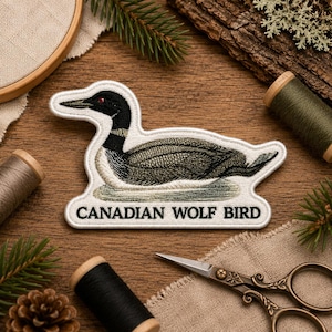 May include: An embroidered patch featuring a loon with a black head, red eye, and grey-brown body, resting on a light green patch of water. The text CANADIAN WOLF BIRD is stitched in black capital letters on a white border at the bottom.