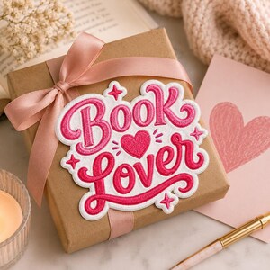May include: A square brown gift box wrapped with a light pink satin ribbon, featuring a white embroidered patch with the hot pink text "BOOK lover" and a small heart icon, accented by four small star shapes on the edges of the patch.