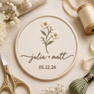 May include: A circular embroidered patch on off-white fabric featuring a floral design in white and green thread. The text "julia + matt" is stitched in dark brown cursive script above the date "05.12.26" in a simple dark brown font.
